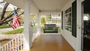 This is a traditional wooden front porch with swinging seat and white pillars. An American flag is displayed in the front yard.