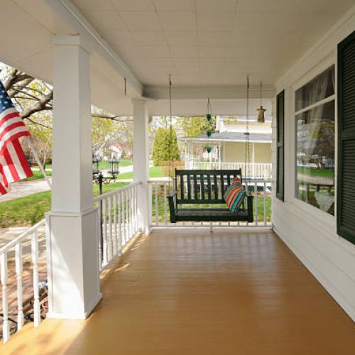 This is a traditional wooden front porch with swinging seat and white pillars. An American flag is displayed in the front yard.